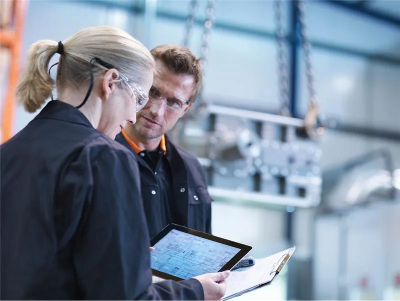 Two engineers in a manufacturing facility reviewing technical plans on a tablet and clipboard, with industrial equipment in the background.